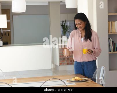 Asian woman standing at dining table holding ceramic mug and orange juice, plate nearby, copy space Stock Photo