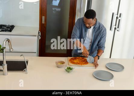 African American man slicing round cheese pizza with pizza cutter on kitchen island, copy space Stock Photo