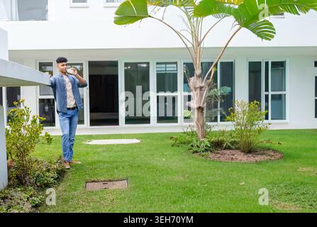 Man standing drinking from bottle, leaning on ledge by palm tree in courtyard, copy space Stock Photo