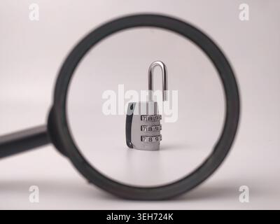 An open silver combination padlock with number dials is framed and scrutinized through a black magnifying glass lens. Stock Photo