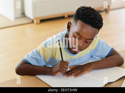 African American boy in striped tee writing in spiral-notebook at wood table at home holding pencil Stock Photo