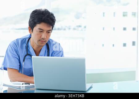 Middle-aged Asian man sitting at clinic desk using laptop while wearing blue scrubs and stethoscope Stock Photo