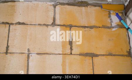 Close up view of paint roller applying yellow primer to masonry block wall. Home improvement or construction process. Worker preparing surface for pai Stock Photo