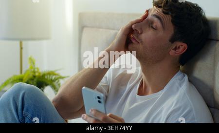 Tired upset Caucasian business man sitting bed in bedroom using mobile phone online stress work exhausted male guy overworked entrepreneur freelancer Stock Photo