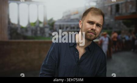 Man with beard tilting head and squinting slightly on an old town street by a stone railing with blurred tourists and buildings; thoughtful. Stock Photo