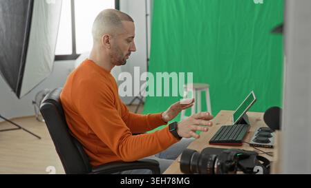 Man photographer pointing at tablet and gesturing with hands at desk in studio, green screen and camera visible; concentration creativity. Stock Photo