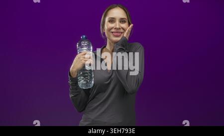 Woman holding a large plastic water bottle and waving right hand in studio with purple backdrop, smiling at camera; hydration joy. Stock Photo