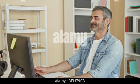 Man typing at computer with hand on keyboard and waving hand while seated in a building office, gesturing to screen; focused remote work. Stock Photo