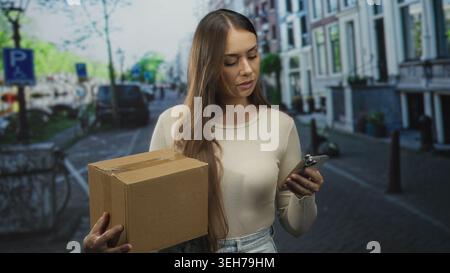 Woman holding cardboard box and smartphone looks at phone while standing on an urban street with parked cars and bicycles; focused delivery. Stock Photo