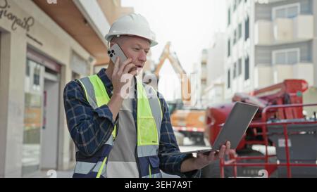 Man builder in hardhat and hi vis vest talks on phone while holding laptop on an urban street with excavator behind him; focus duty. Stock Photo
