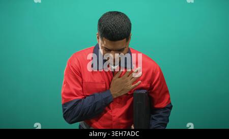Hispanic bearded man in red work uniform stands holding a toolbox with one hand and places his hand on chest in a studio setting; pride duty. Stock Photo