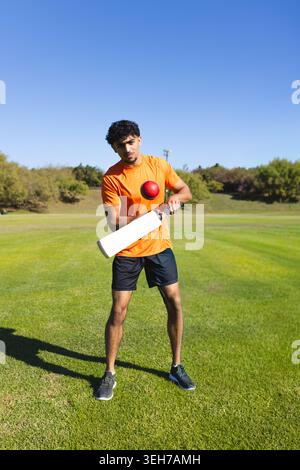 Indian man in orange shirt black shorts standing on field holding white bat and red ball Stock Photo