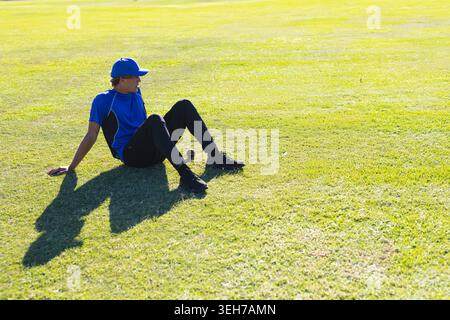 Male athlete sitting on grassy field wearing blue shirt and cap near baseball, casting shadow Stock Photo