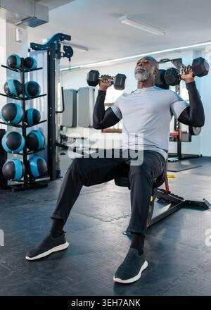 Senior African American man in activewear doing shoulder press at gym bench holding dumbbells Stock Photo