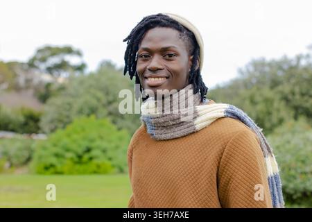 African American man standing and smiling in park wearing cream beanie, knit scarf, brown sweater Stock Photo