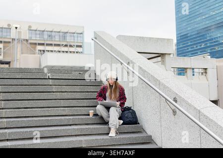 Woman sitting on concrete plaza stairs wearing beanie and headphones using laptop, copy space Stock Photo