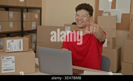 Man in red shirt with laptop points finger toward camera amid stacked shipping boxes and parcels on shelves in building; entrepreneurial pride. Stock Photo