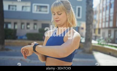 Woman checks smartwatch on wrist while standing on a sunlit urban street near apartment building in blue sportswear; training focus. Stock Photo