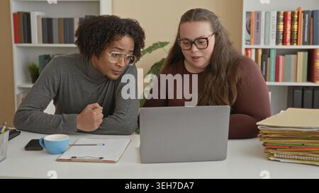 Man and woman leaning over laptop at desk with clipboard, coffee cup and stacked folders, man making a fist gesture while studying the screen in a bui Stock Photo