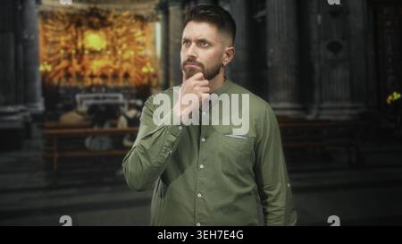 Hispanic man with beard stands with hands behind back and hand to chin gesture inside ornate church building; quiet contemplation. Stock Photo