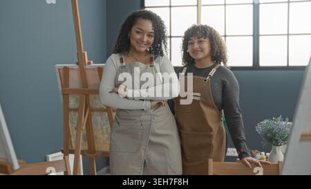 Woman wearing apron with arms crossed standing beside easel and canvas in art studio; creative friendship. Stock Photo