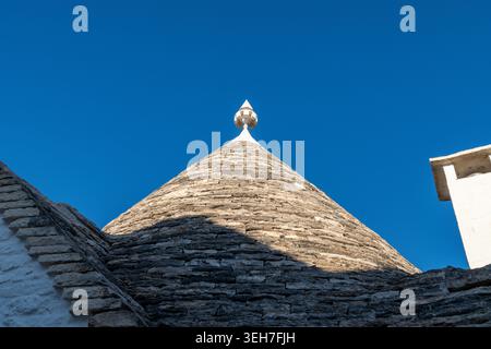 A detailed view of a traditional conical stone roof with white finial against a deep blue sky, captured in the town of Alberobello in Italy. Stock Photo