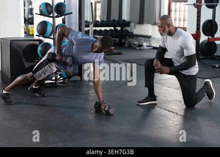 African American male trainer and client doing side-plank row in gym with hex dumbbell, copy space. Weights, coaching, focus, fitness, equipment, daylight, strength Stock Photo