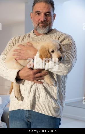 Senior man standing in living room holding golden retriever puppy, wearing cream cable-knit sweater. Dog, companion, home, soft, mature, cozy, pet Stock Photo