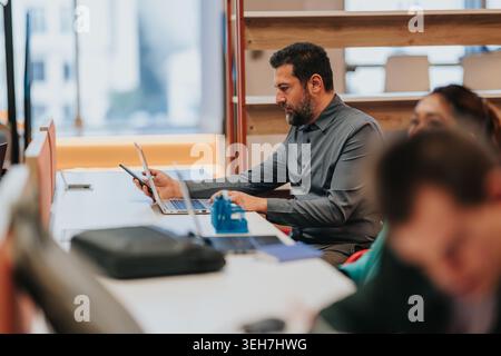 Businessman working on laptop and smartphone at shared office desk with coworkers nearby Stock Photo