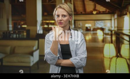 Woman extends hand palm up and beckons in a building lobby, arm crossed at waist; demanding expectation. Stock Photo