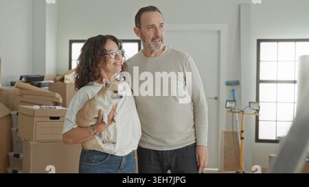Man pointing finger while embracing woman holding small dog amid moving boxes in building; hopeful new beginning. Stock Photo