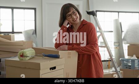 Woman leaning on cardboard box with paint roller nearby, hand on cheek in building; fresh start hopeful. Stock Photo