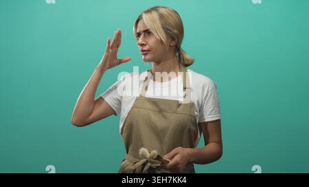 Woman makeup artist young blonde wearing apron pinches nose while holding brush in studio with teal backdrop; disgust reaction. Stock Photo