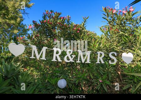 A white MR and MRS banner with hearts hangs against lush green bushes with pink flowers and a clear blue sky, creating a romantic wedding decoration b Stock Photo