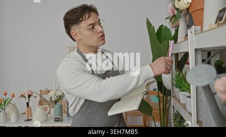 Man florist writes in notebook with hand holding pink pen beside potted plants in studio; quiet focus. Stock Photo
