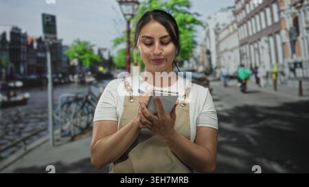 Young woman waitress writing on a notepad with pen on an amsterdam street canal side, wearing apron while taking an order; service concentration. Stock Photo
