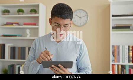 Man holding tablet and stylus, writing on touchscreen and glancing upward in a study, wearing glasses and sweater with books and clock visible; focuse Stock Photo