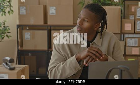 Man with packages clasping hands over laptop in a storage building with stacked boxes and scanner visible; small business pride calm. Stock Photo