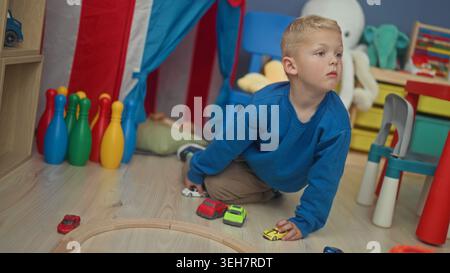 Boy kneeling on floor pushing toy car with right hand near wooden track and colorful bowling pins in building; curiosity imagination. Stock Photo