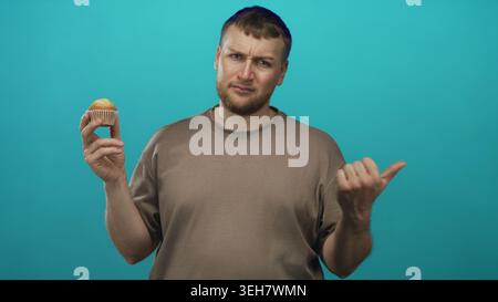 Man holding muffin with thumb pointing sideways in studio with teal backdrop, young heavyset wearing brown tee and puzzled frown; doubt hesitation. Stock Photo