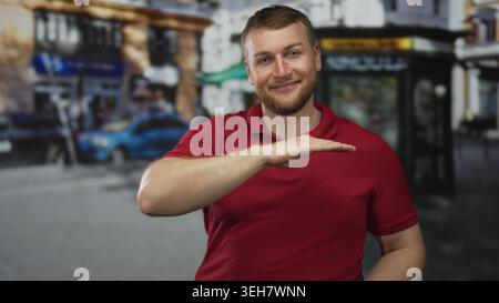 Man in red polo holds hands level as if framing an invisible box on street near kiosk and parked car; confidence ease. Stock Photo