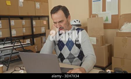 Middle aged man at laptop with hand on cheek among stacked cardboard boxes in a building; small business stress. Stock Photo