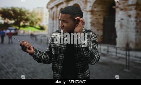 Young black man holding screwdriver and cupping ear on a cobblestone street near an ancient stone facade; confusion seeking help. Stock Photo