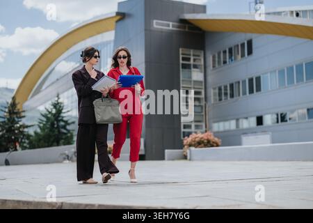 Two businesswomen walking and discussing documents outside a modern office building Stock Photo