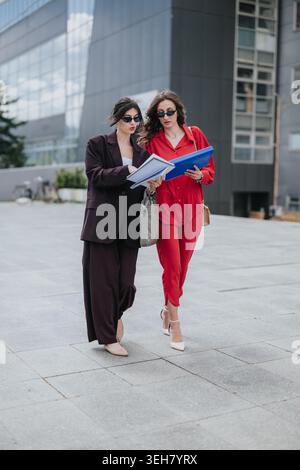 Two businesswomen walking and reviewing documents outside a modern office building Stock Photo