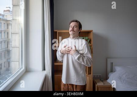 Focused man practicing mindful breathing, feeling heartbeat, hands on chest, eyes closed at home.  Stock Photo