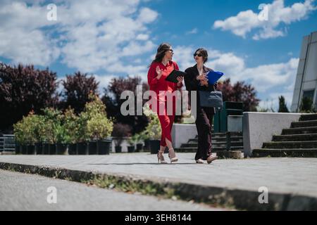 Two businesswomen walking and discussing documents on a sunny urban sidewalk during a casual meeting Stock Photo