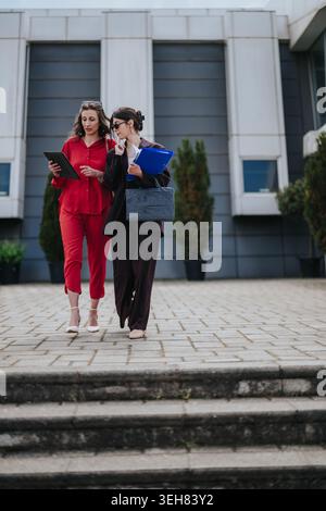 Two businesswomen walking and discussing documents outside a modern office building Stock Photo