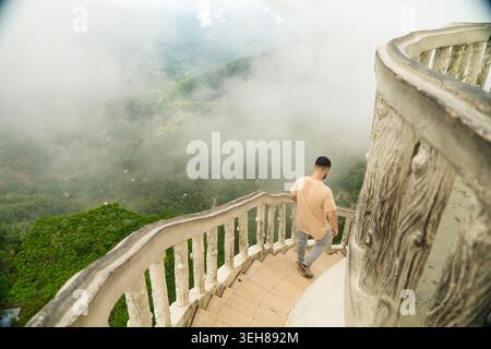 Sri Lanka - 22 February 2026: View of a man walking down a spiral staircase with white railings overlooking a lush green valley covered in thick fog. Stock Photo