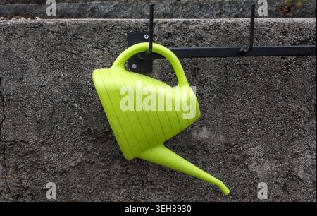 Bright green plastic watering can hanging on a metal hook against a textured grey concrete wall backdrop. Stock Photo